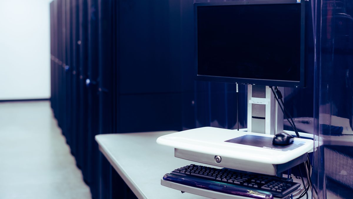 Contemporary computer with black screen placed on stand near row of server steel racks in data center.