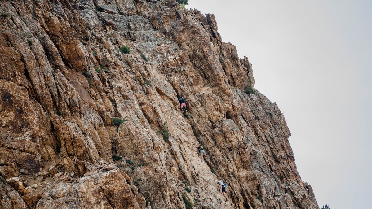 Three climbers spaced out on a steep, sunlit limestone cliff under a pale grey sky, each on a separate line up the same rock face.