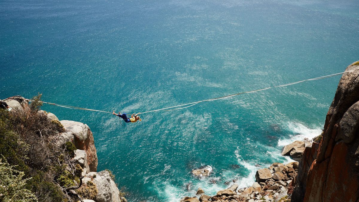 High angle of unrecognizable female tourist on zipline between rocky cliffs over turquoise rippling sea water washing stony shore.