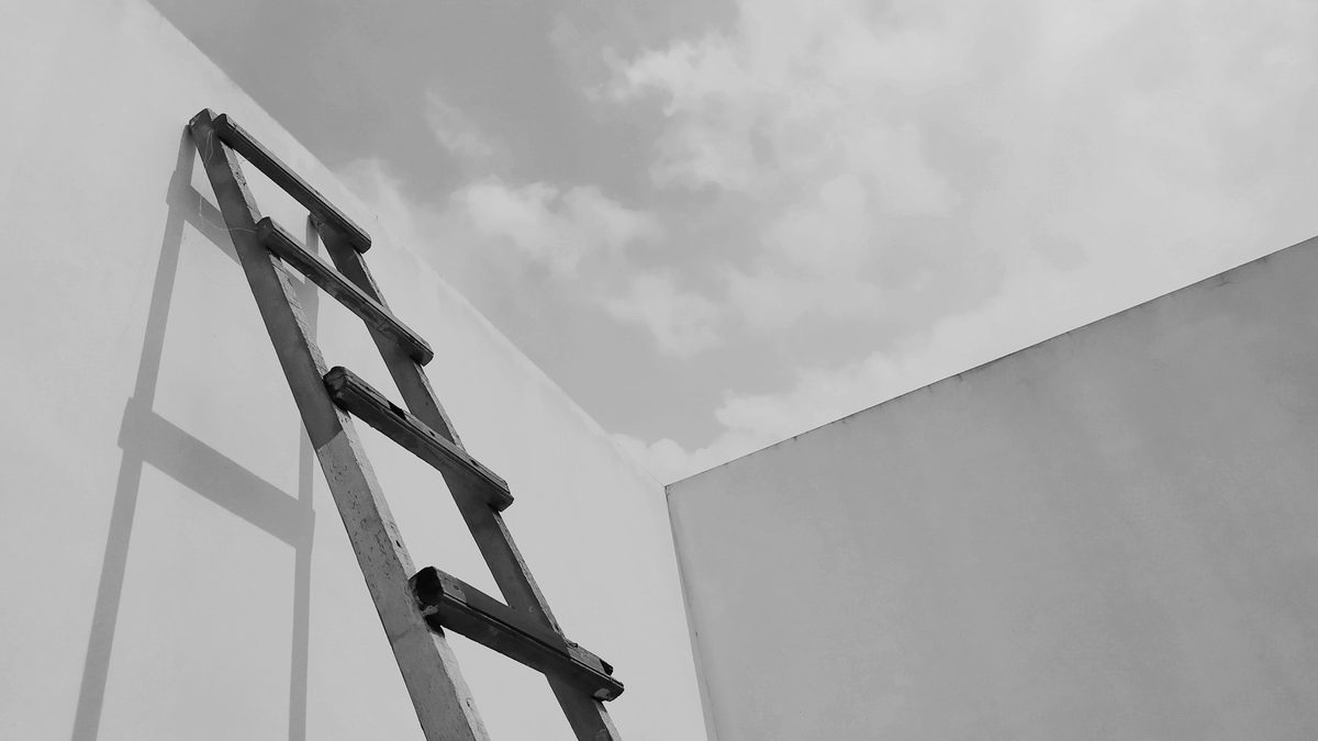 Simple black and white image of a ladder leaning against a white wall under a cloudy sky.