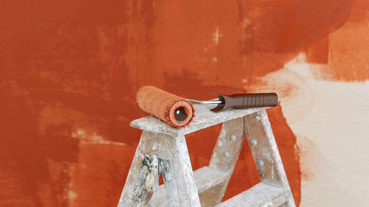 Paint roller on a ladder with a partially painted red wall in the background.