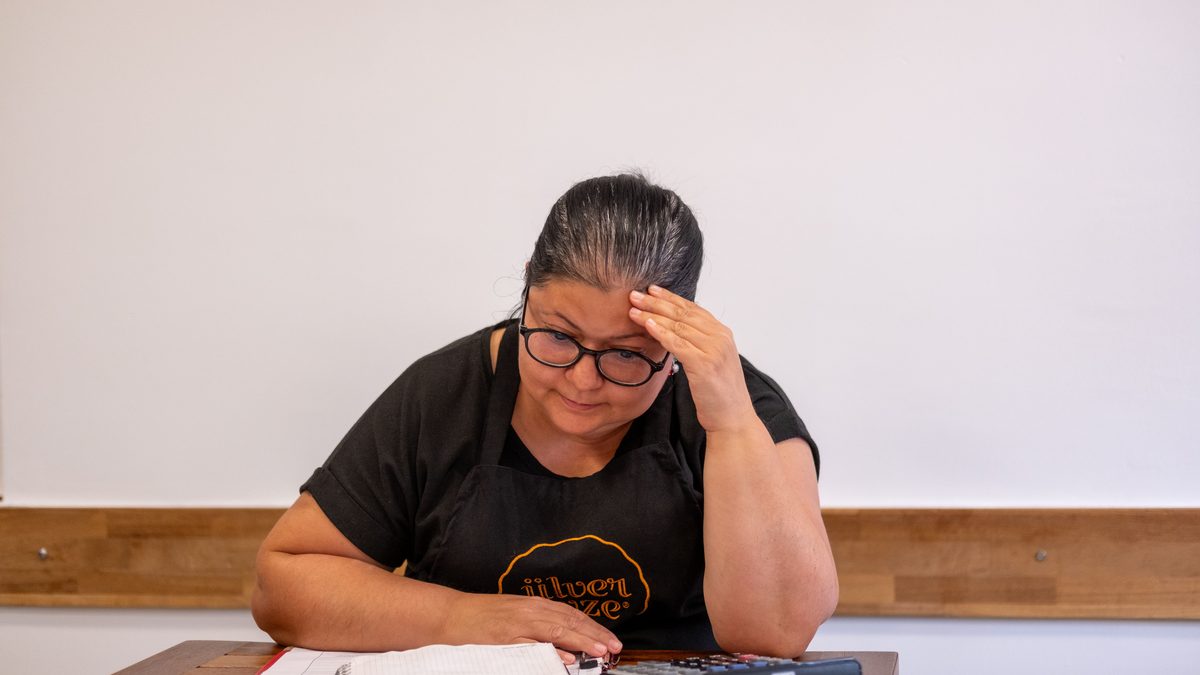 Photograph of a woman at a desk looking strained as she works through household finances with a calculator, a pen and handwritten notes in front of her.