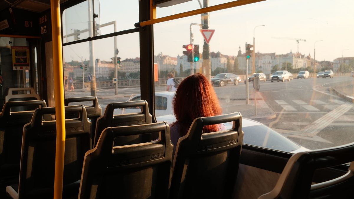 A passenger with long red hair sits alone on a near empty bus, looking out of the window at a road junction in low evening sunlight.