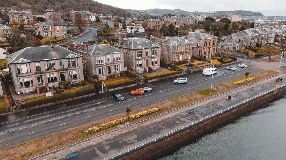 A scenic aerial view of Greenock, Scotland featuring Victorian homes along a coastal road with visible traffic.