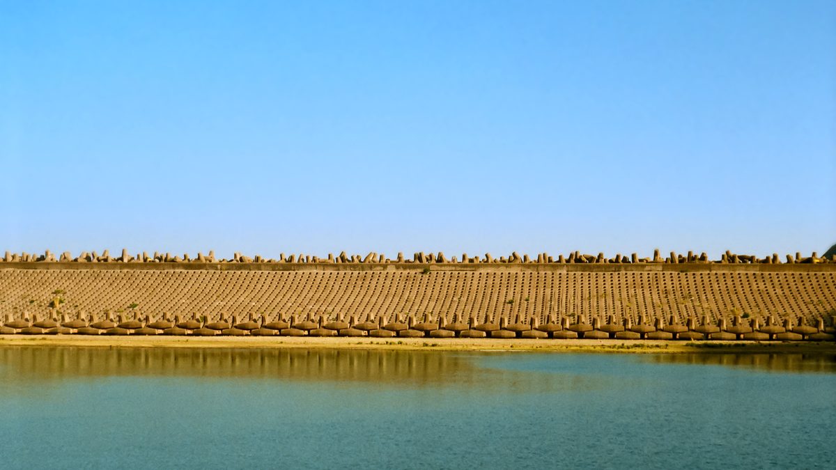 A still reservoir held behind a long embankment lined with hundreds of concrete wave-breaking blocks, under a clear blue sky.