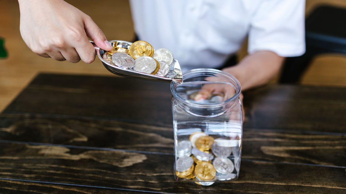 Hand placing gold and silver coins into a glass jar on a wooden table indoors.