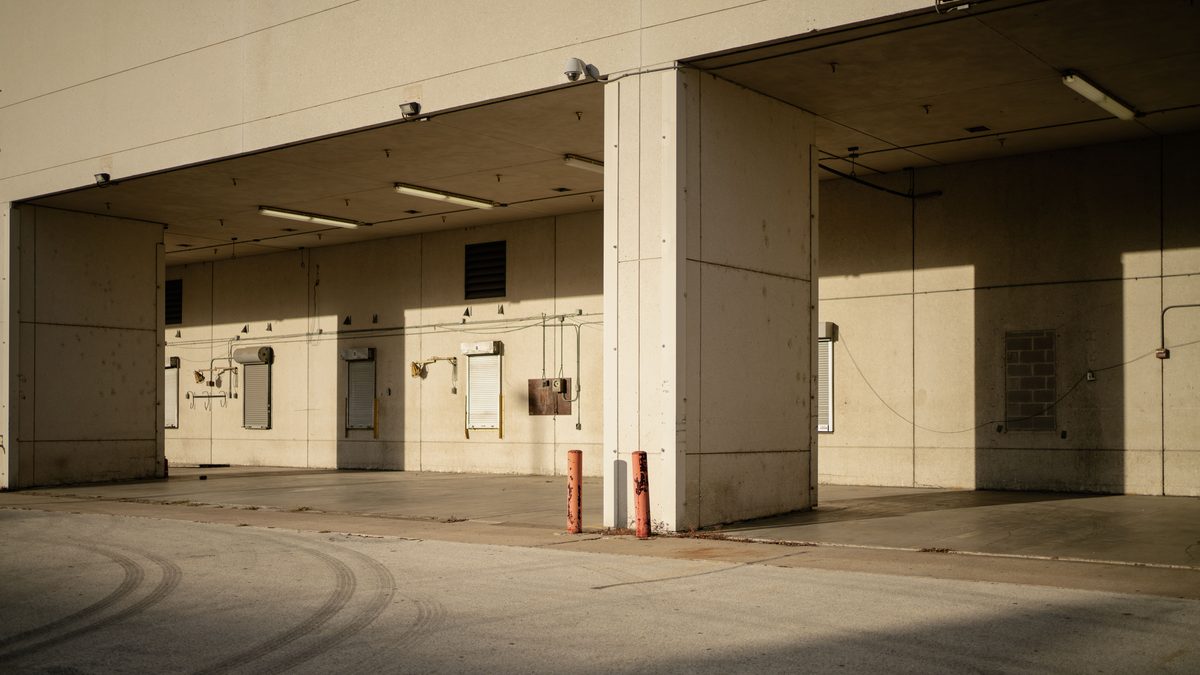 A row of covered loading bays on an industrial unit with the shutters down, low sun throwing hard shadows across the concrete apron and two red bollards.