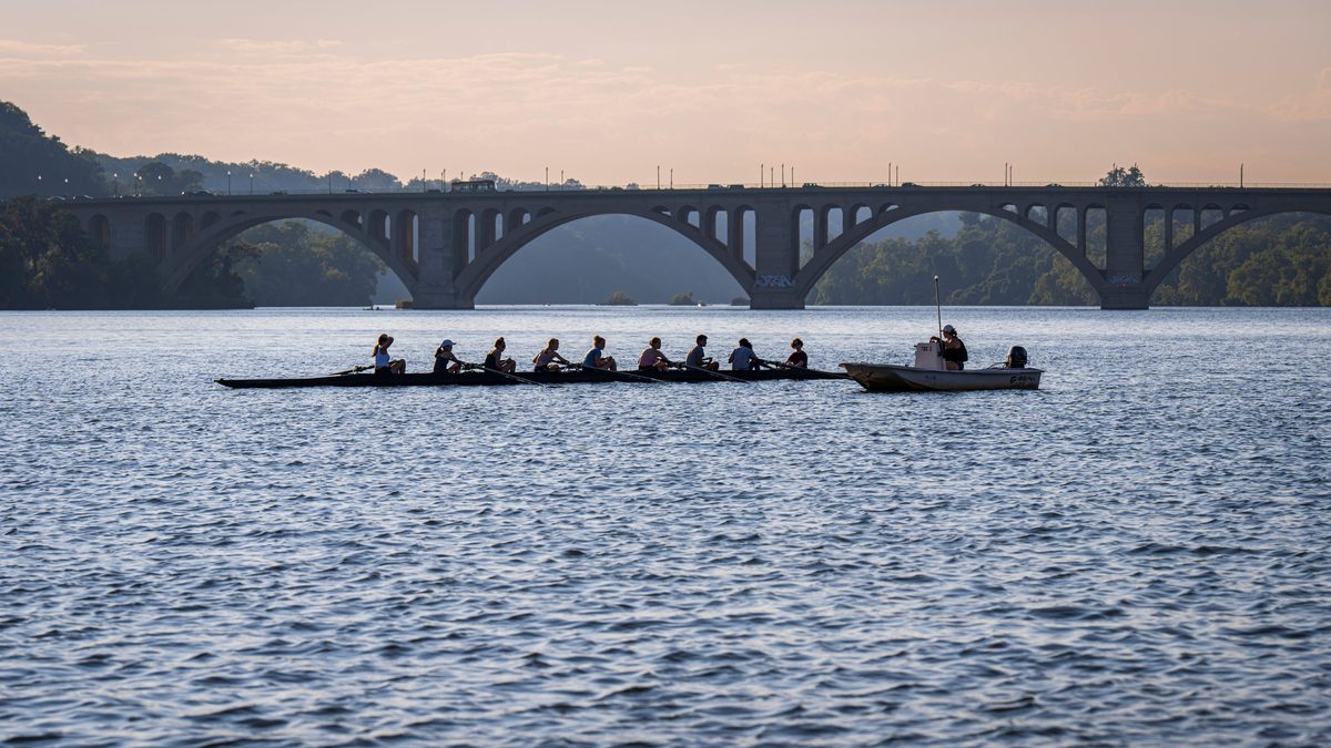 An eight-oared rowing shell crossing wide open water at dusk, the crew silhouetted against a pale sky, with a coach's launch alongside and a long stone arch bridge and wooded bank behind.
