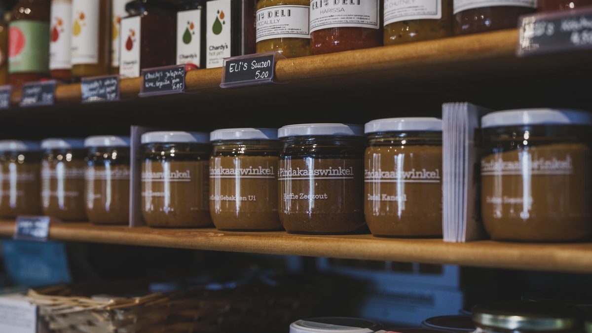 A row of identical brown glass jars with white lids lined up along a wooden shop shelf, beneath a higher shelf of assorted sauce and chutney bottles of different shapes and sizes.