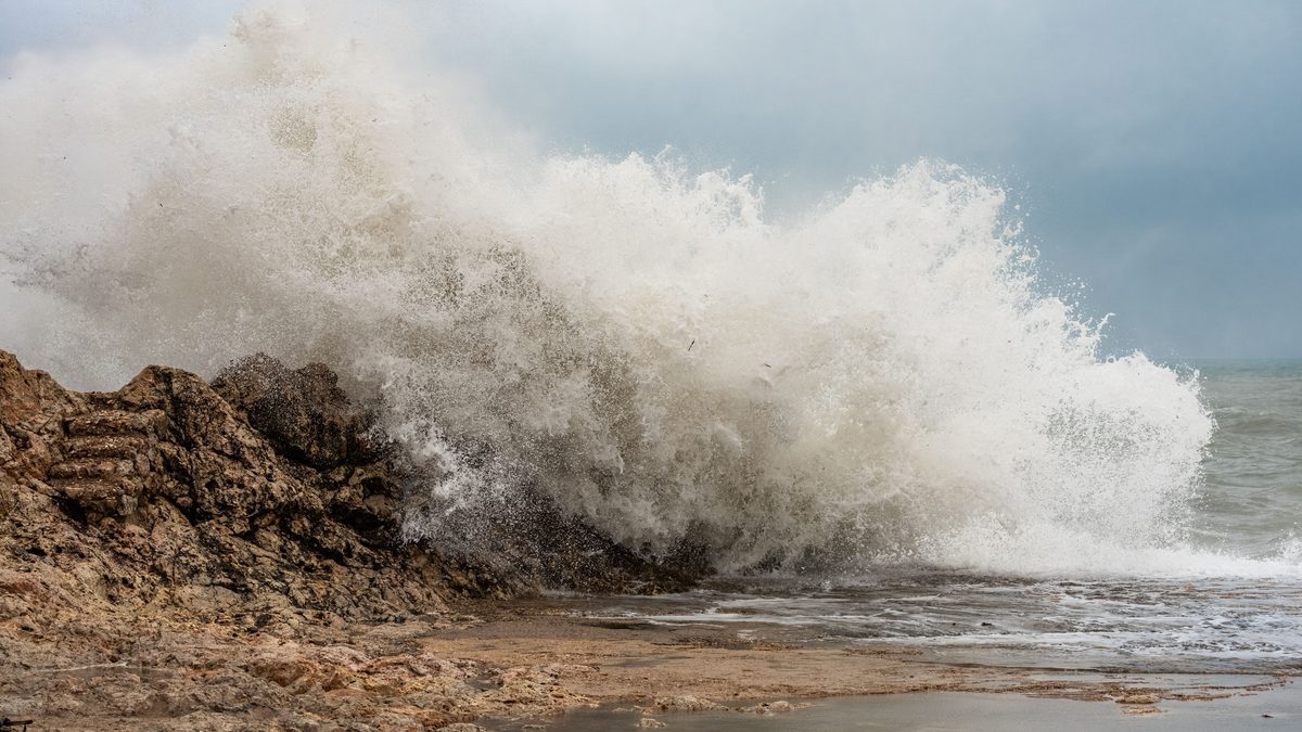 A large white-water wave breaking over a low rocky headland on a grey coast, spray thrown high above the rocks while calmer shallow water lies on the sand in the foreground.