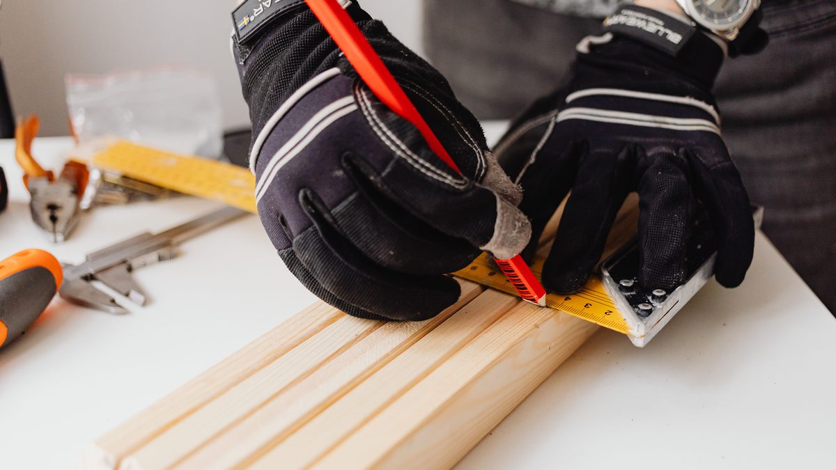 Close-up of gloved hands using ruler to measure wood in a carpentry workshop. Tools are visible in the background.