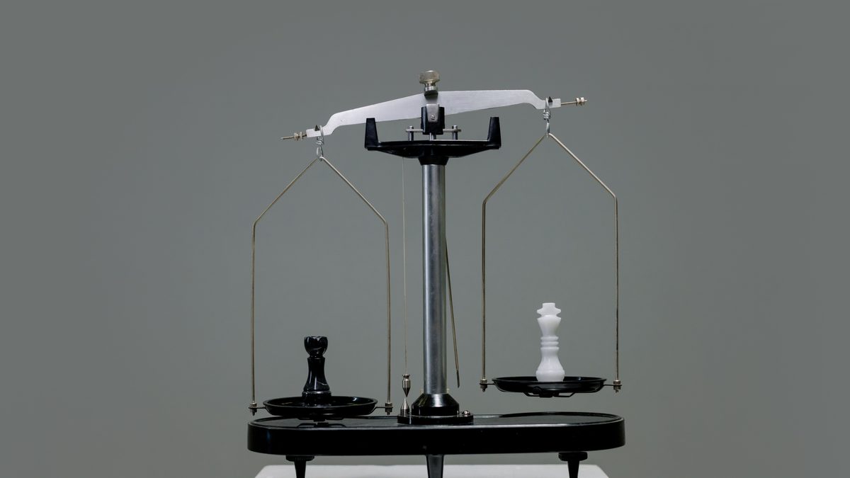 A small two-pan balance scale on a grey background, tipped down to the left, with a black chess pawn sitting on the lowered pan and a white chess queen on the raised pan opposite.