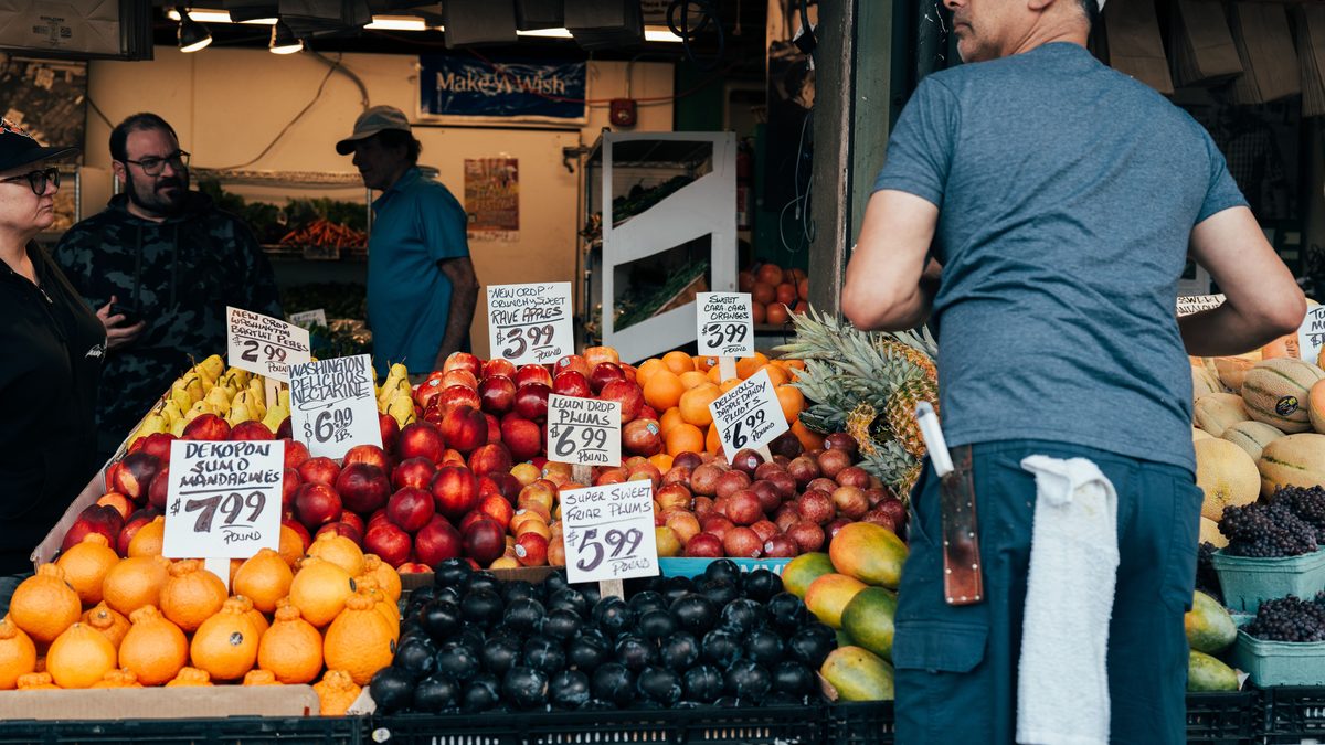 An outdoor fruit stall with mandarins, nectarines, plums and oranges in crates, each variety fronted by a large handwritten card giving its price per pound, while a shopkeeper in a grey t-shirt stands at the right.
