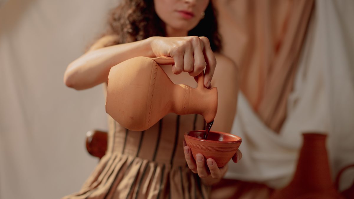 Elegant woman in dress pouring liquid from a clay jug into a bowl. Warm tones.