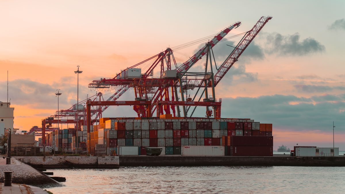 Rows of container gantry cranes standing over stacked shipping containers at a port, silhouetted against an orange sunset.