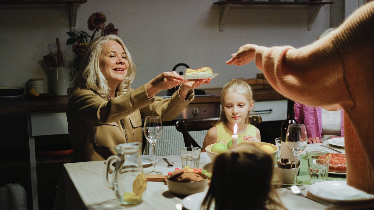 An older woman reaches across a candlelit family dinner table to pass a plate of cake to a young girl, with glasses, bowls and another adult's outstretched arm in the foreground.