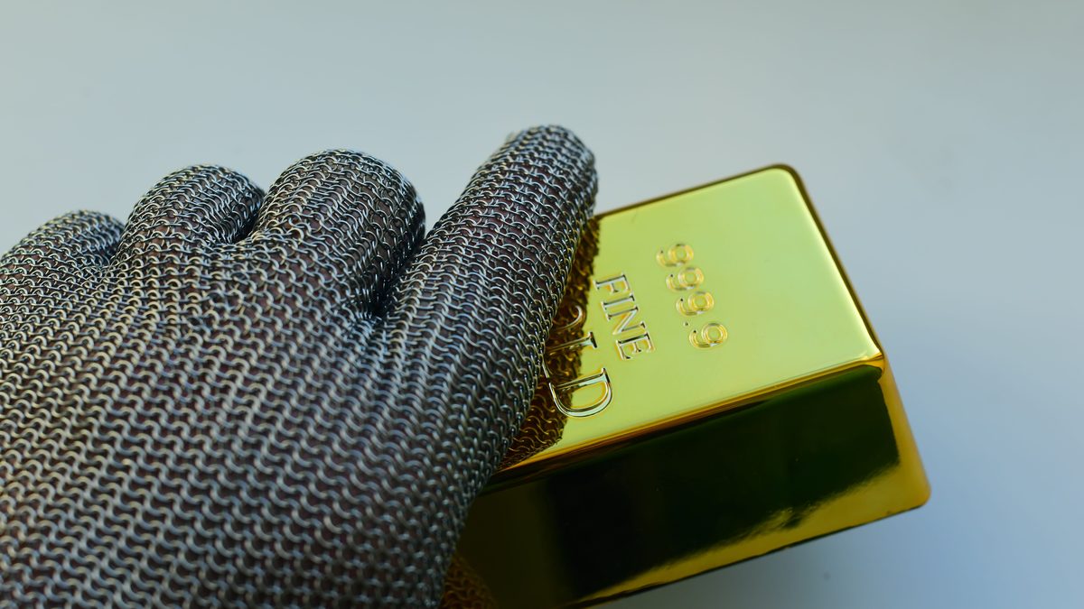 Close-up of a metal gloved hand holding a shiny gold bar indoors.