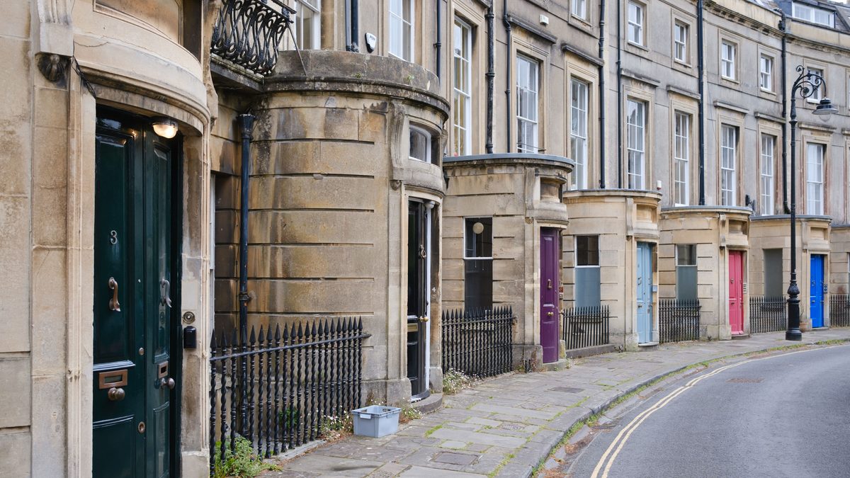 A curved row of Georgian terraced houses in Bath, each with a brightly painted front door, on an empty pavement under flat daylight.