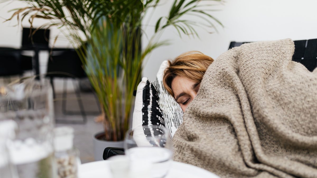 A woman resting on a couch covered by a blanket, indoors with plants.