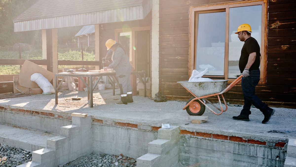 Two construction workers on a half-built house in low sun, one pushing an orange wheelbarrow of materials across a concrete slab while the other works at a trestle bench.