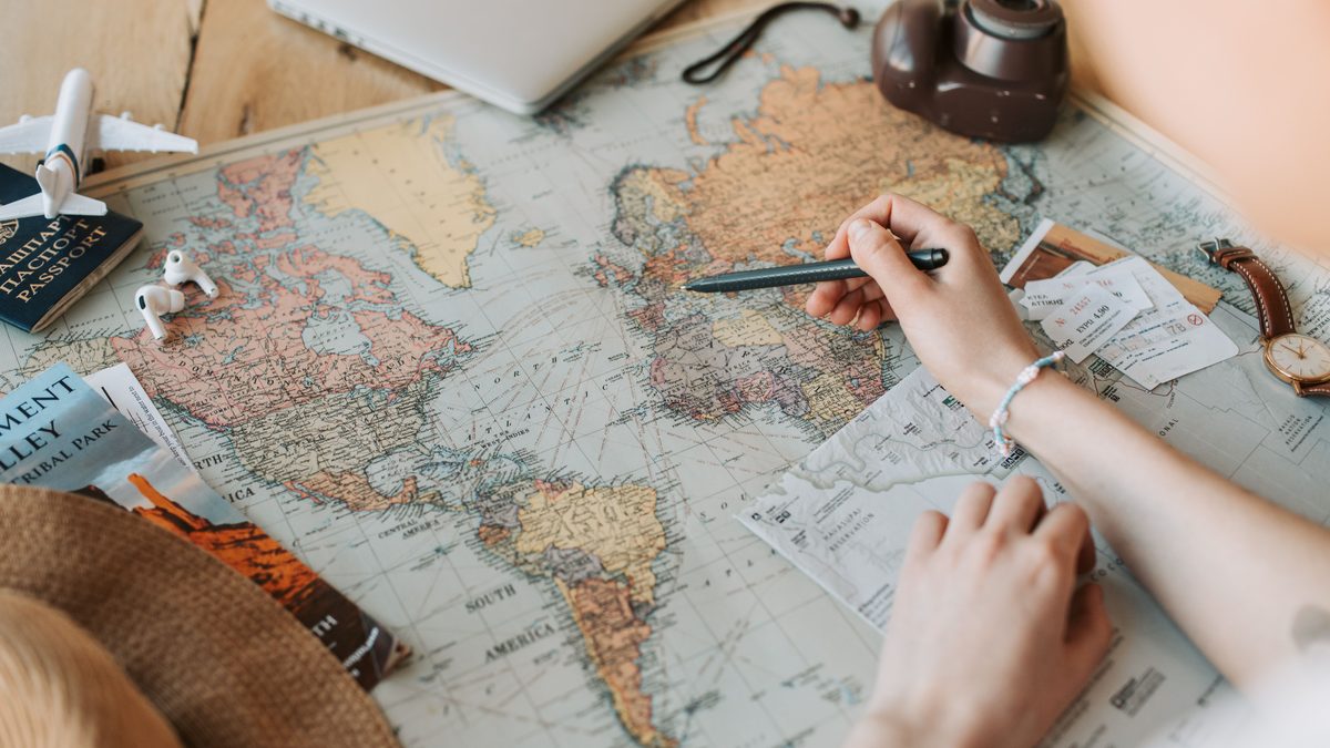 Hands marking destinations on a world map surrounded by travel essentials like passport and camera.