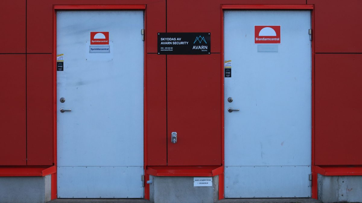 Two white doors against a red wall featuring security signs and logos.