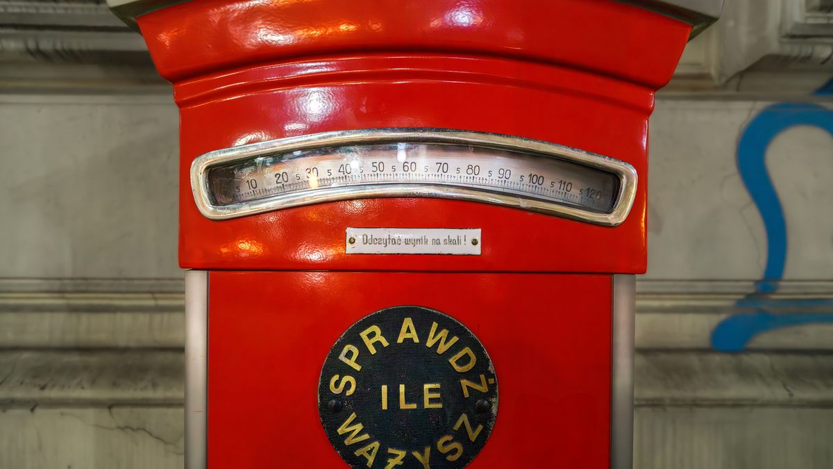 Close-up of a vintage red coin-operated weighing machine, its curved dial marked from 5 to 120 and a Polish plate reading Sprawdz ile wazysz.