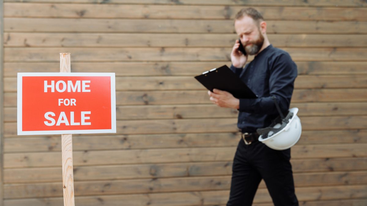Real estate agent on phone with 'Home for Sale' sign outside a property.