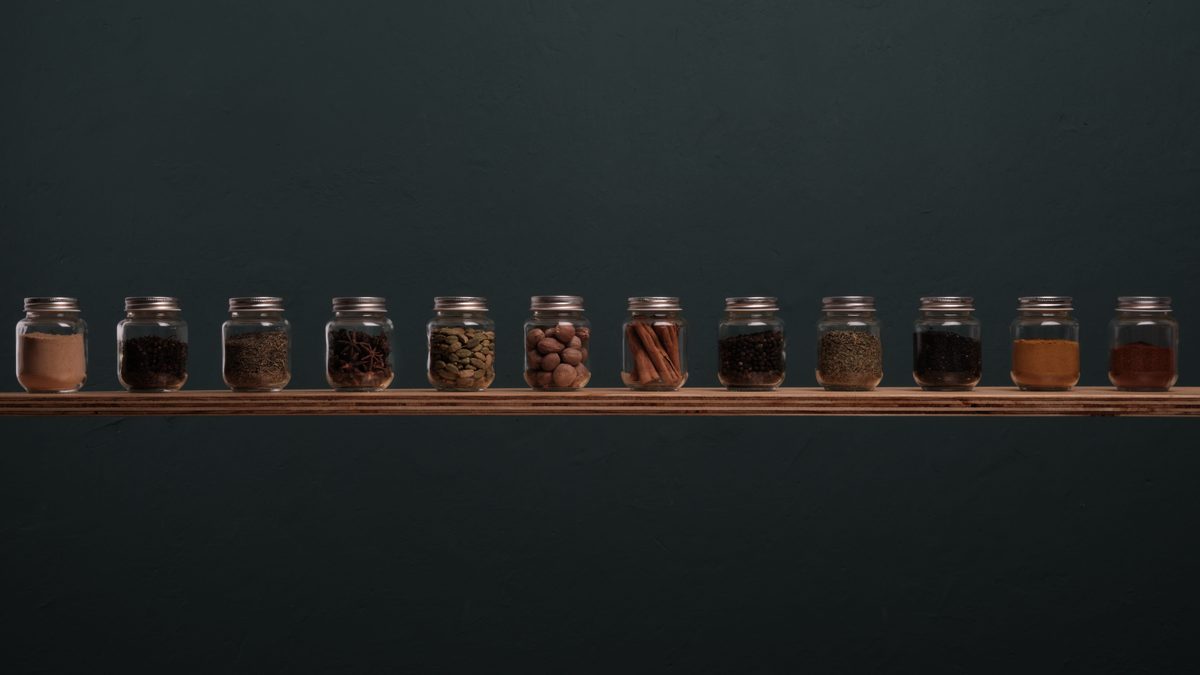 A row of identical glass spice jars standing on a narrow wooden shelf against a dark green wall, each jar filled to about the same level with a different spice.