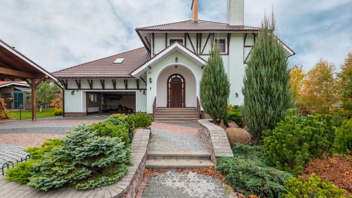 A detached house behind a wide block-paved forecourt, with an open garage to one side, clipped conifers in the beds and a stone-set path climbing to the front door.