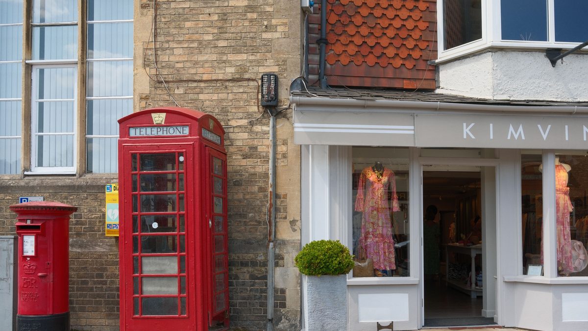 A red Royal Mail pillar box and a red telephone box standing on a British high street, in front of a brick building and an open boutique shopfront.