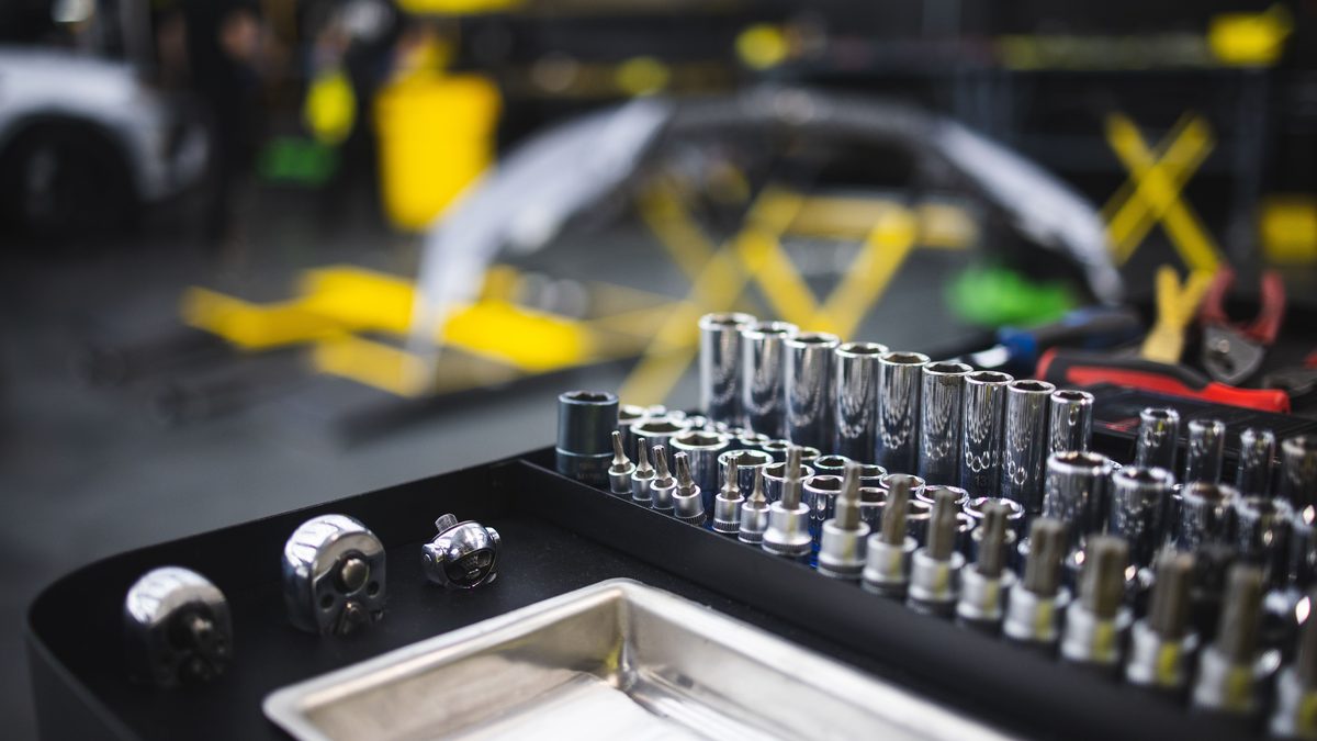 A tray of chrome sockets and driver bits laid out in graduated sizes, with three ratchet heads on the bench beside them and a blurred workshop and yellow lift behind.