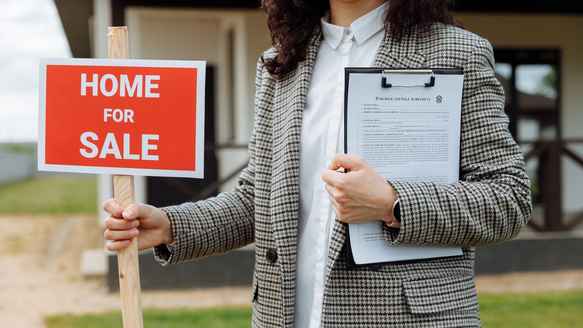 A real estate agent holding a home for sale sign and clipboard outside a property.