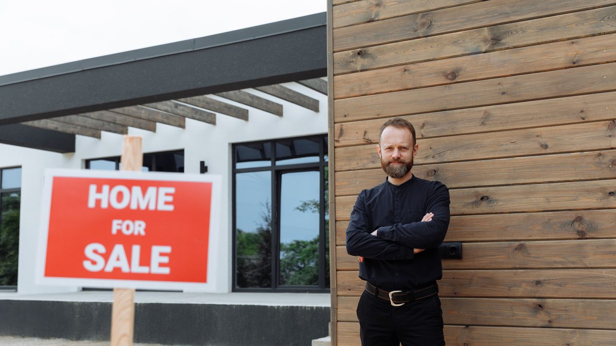 Real estate agent standing by a modern home with a 'Home for Sale' sign.