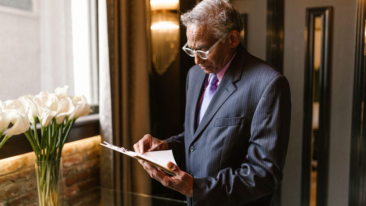 Senior man in a suit reading papers carefully by a window. Indoor setting with elegant decor.