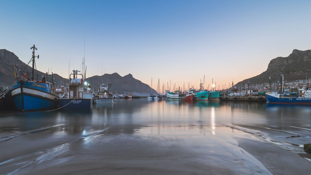 Captured at sunset, this tranquil scene showcases fishing boats reflecting on the calm waters of Hout Bay Harbor.