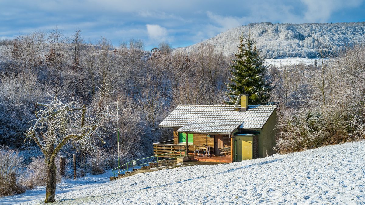 A small wooden holiday chalet standing empty on a snow-covered hillside, its terrace chairs unused and the wooded valley behind it under frost.