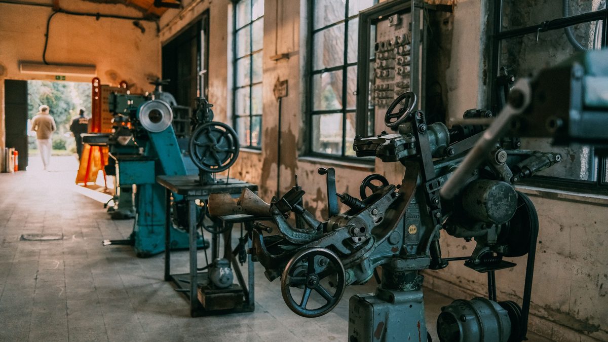 A row of old cast-iron machine tools standing along the wall of a disused workshop, tall grimy windows to one side and a figure walking out through the far doorway.
