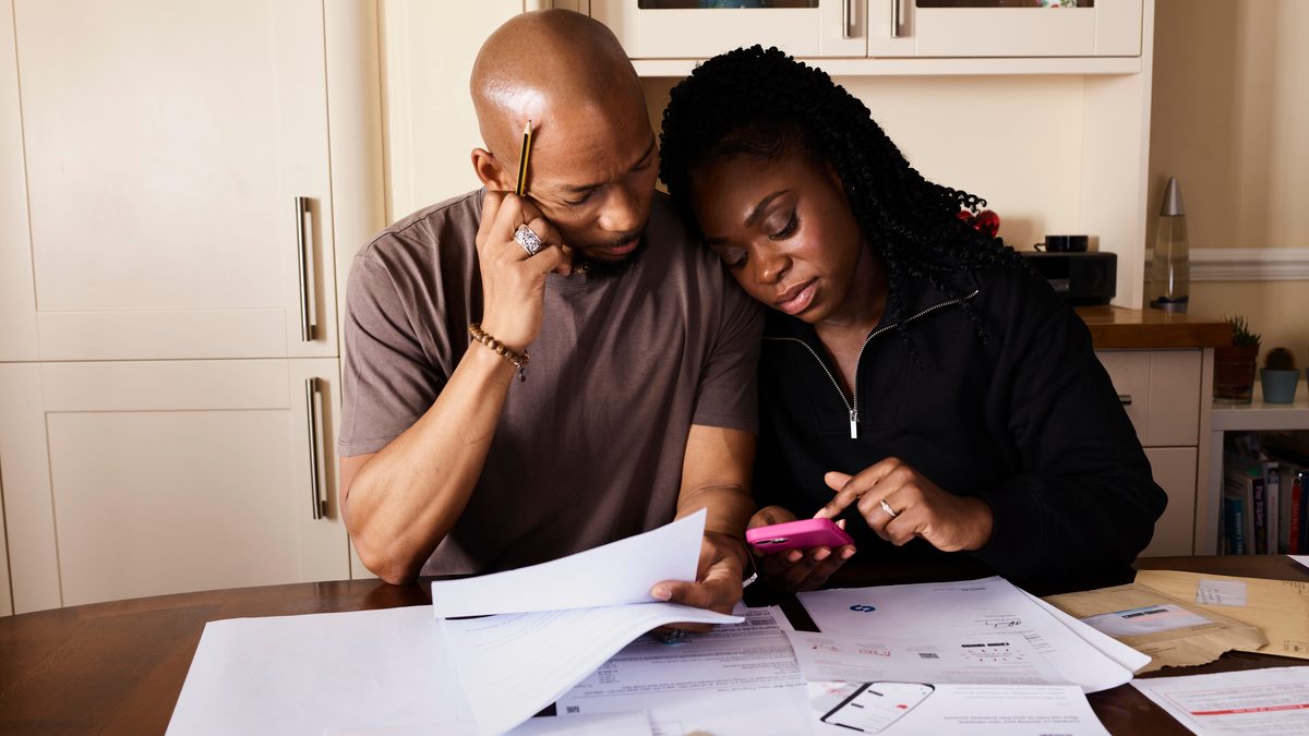 Two people sit close together at a kitchen table covered in loose statements and opened envelopes, one holding a sheaf of papers and the other tapping figures into a phone.