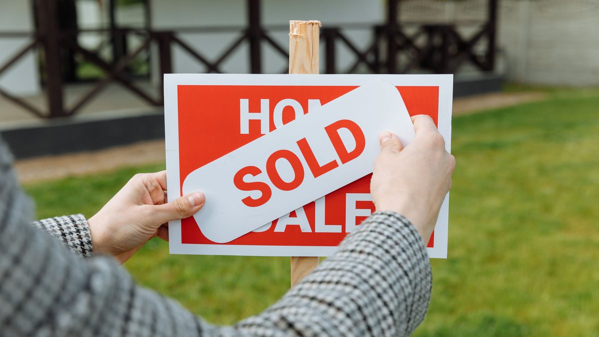 Close-up of hands placing a sold sticker on a real estate sign outside a house.