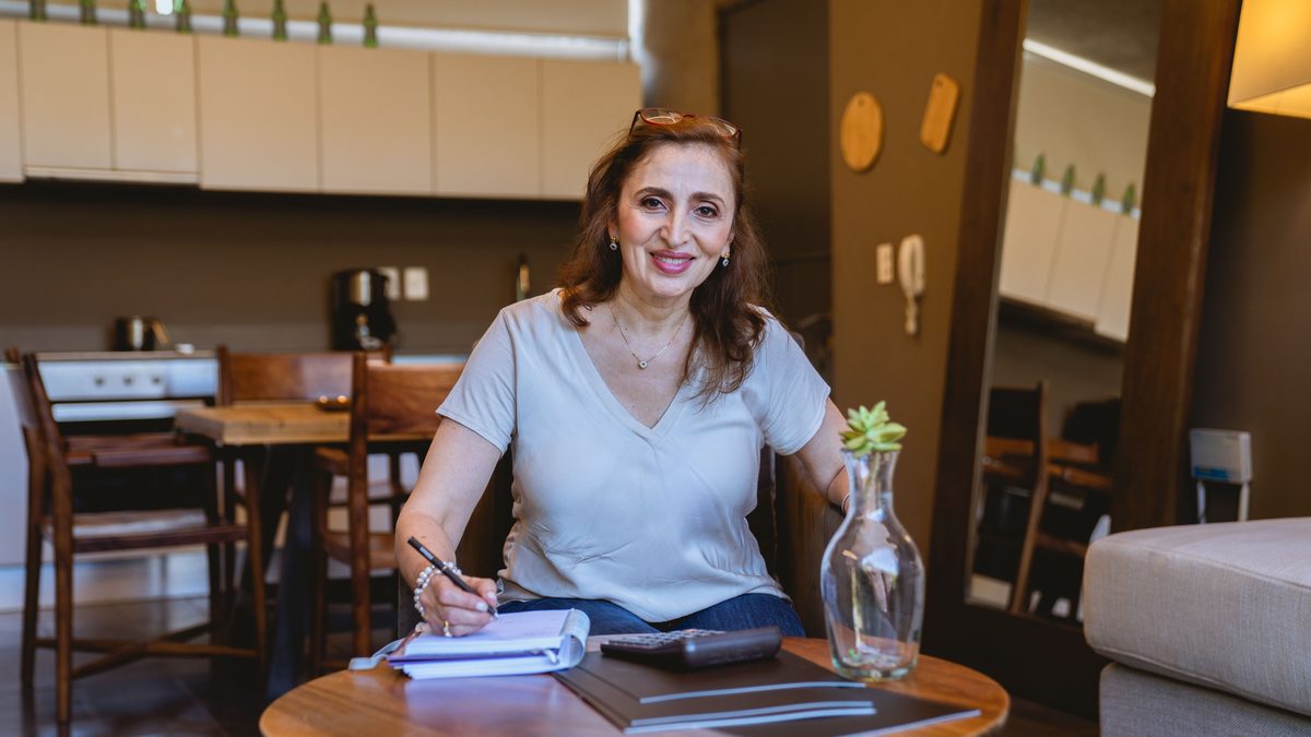 Elderly woman smiling while writing in a notebook, seated in a warm, cozy kitchen.