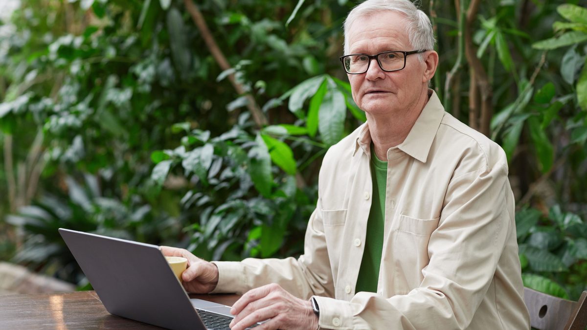 Elderly man using laptop outdoors, surrounded by greenery, remote work concept.