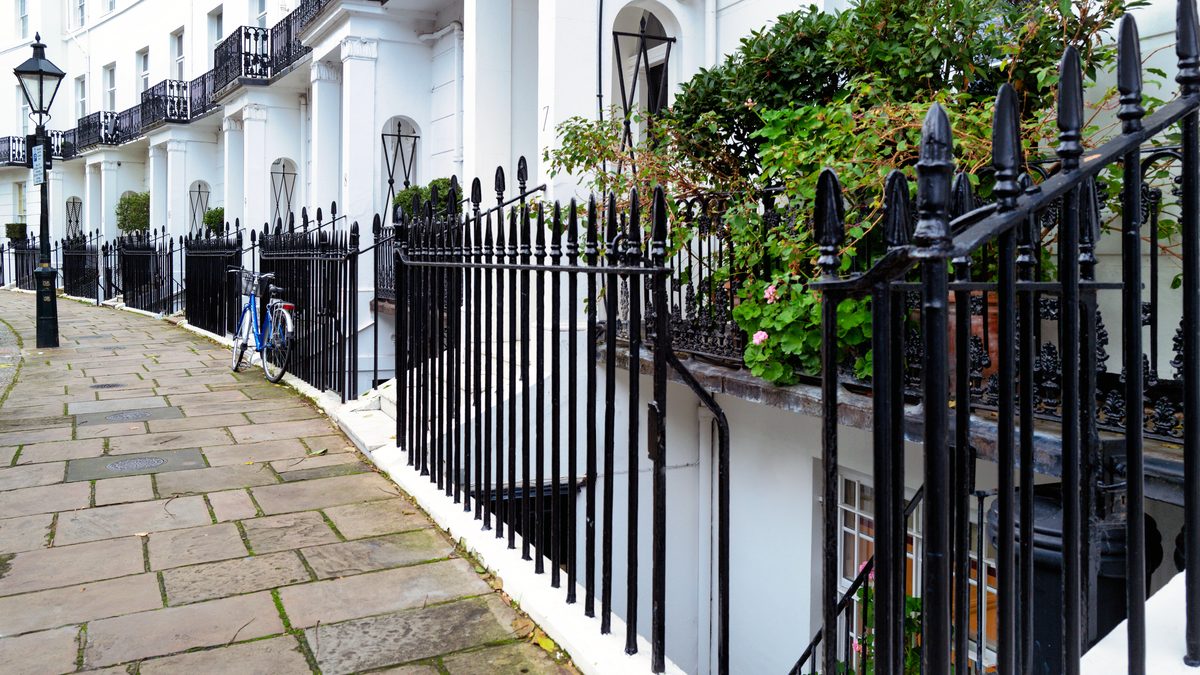 A terrace of white stucco Georgian townhouses with black iron railings and columned porches in Kensington, London, photographed from the pavement in daylight.