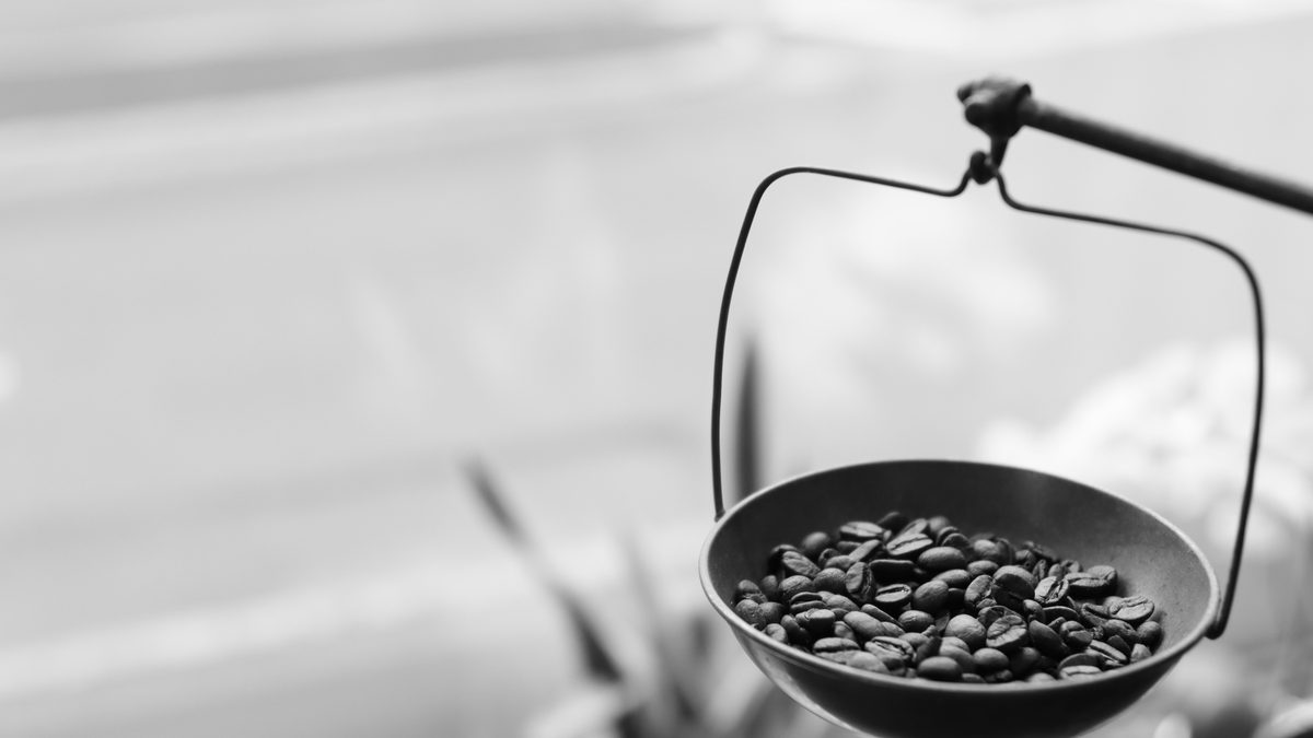 Black and white close-up of a single shallow metal pan of a hanging balance scale, filled with roasted coffee beans, suspended on thin wire against a soft pale background.
