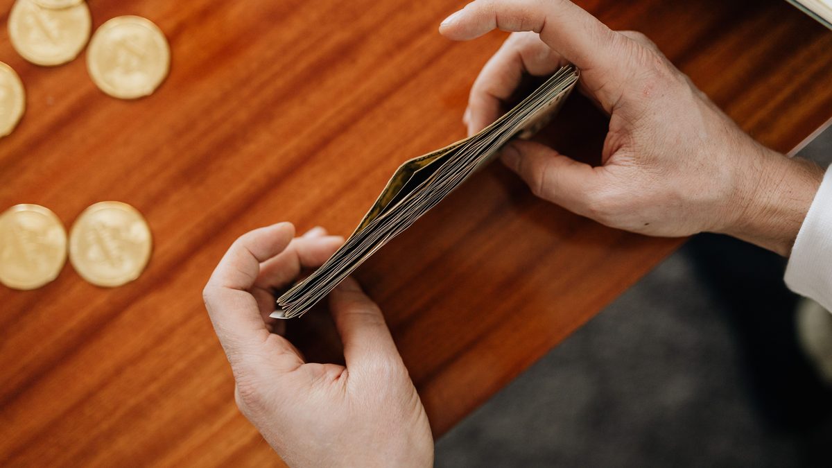 Two hands fanning through a folded stack of banknotes on a polished wooden desk, with gold-coloured coins laid out to the left of the frame.